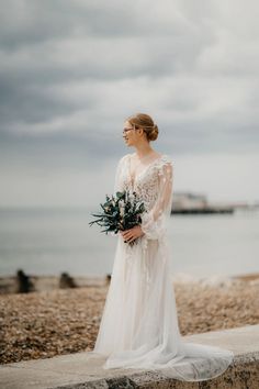 a woman in a wedding dress standing on the beach holding a bouquet of flowers and looking off into the distance