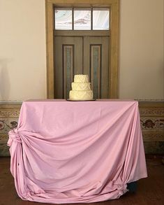 a table covered with a pink cloth in front of a door and a white cake on top of it