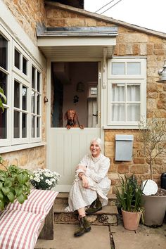 an older woman sitting on the front steps of a house with her dog in the window