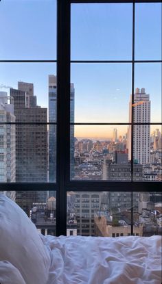 a bed with white sheets and pillows in front of a large window overlooking the city