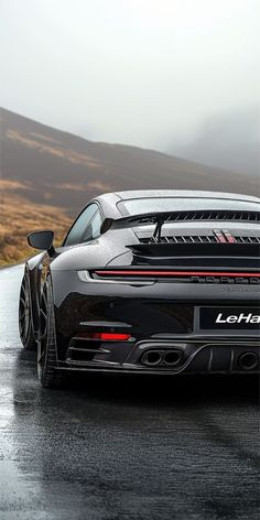 the rear end of a black sports car on a wet road with mountains in the background