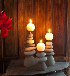 three lit candles sitting on top of stacked rocks in front of a wooden wall with red flowers