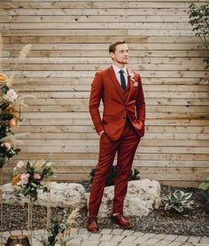 a man in a red suit and tie standing next to a wooden wall with flowers