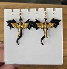 These handmade earrings feature a large black dragon and a smaller gold dragon, inspired by the book Fourth Wing. The dragons reference Tairn and Andarna from the book! These are made of polymer clay and are painted (the gold is metallic!) and coated in a nice shiny gloss. The ear studs and rings are golden stainless steel.  You can add a bonus pair of lightening bolt earrings for only $5. Pick between black or white. These reference Violet's lightening power. These are handmade and hand painted Fourth Wing Jewelry, Dragon Earrings, Wolf Jewelry, Gold Dragon, Magical Jewelry, Bookish Gifts, Fantasy Jewelry, Girly Jewelry, Fun Earrings