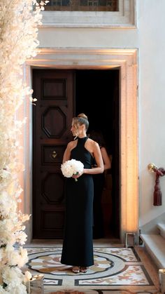 a woman standing in front of a doorway holding a bouquet of flowers and wearing a black dress