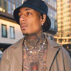 a man with tattoos on his chest wearing a tan jacket and black hat is looking off to the side