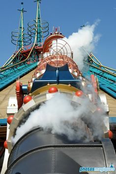 steam billows out from the top of a train at an amusement park with rides in the background