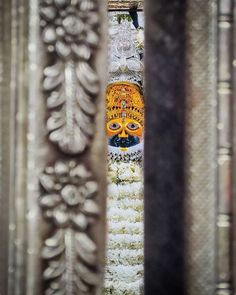 an intricately decorated face is seen through the holes in some metal structures, with white and yellow decorations