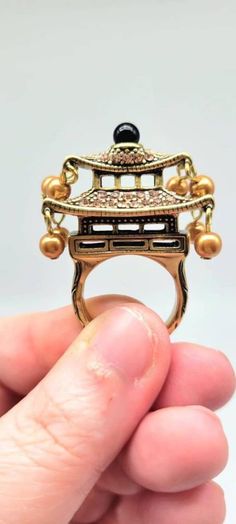 "Chinese Ring, Asian Ring, Temple Ring, Pagoda Ring, Dynasty Jewelry, Pearl Dangle Ring, Chinoiserie Jewelry, Heidi Daus Ring, Chinese Style Measurements: 2-1/8\" L x 1-1/8\" W x 1-5/8\" H Metal: Bronze Tone Finish: Polished, Oxidized, Textured Representing wisdom, peace and harmony, this Chinese temple is as stylish as it is symbolic. Featuring an intricately designed pagoda that is COMPLETELY ENCRUSTED IN SWAROVSKI CRYSTALS and glass pearl drops, tons of beautiful movement and magnificent sparkle await! Shades of stone include champagne diamond crystal, Golden glass pearl and a single jet-black glass bead on top. This piece is so unique in that it has a THREE-DIMENSIONAL look when worn. It sits a full inch off of your finger and can be seen from every single angle! A great addition to an Antique Chinese Jewelry, Chinese Character Ring, Chinoiserie Art Deco, Chinese Jewelry, Heidi Daus, Champagne Diamond, Diamond Crystal, Pearl Drop, Fun Earrings