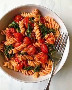 pasta with tomatoes and spinach in a white bowl next to a fork on a marble surface