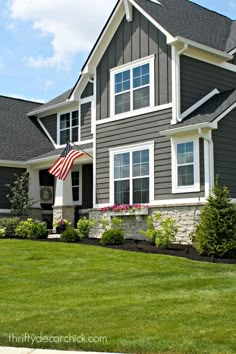 an american flag is in front of a gray house with green grass and flowers on the lawn