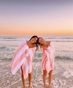 two women in pink and white towels standing on the beach with their arms around each other