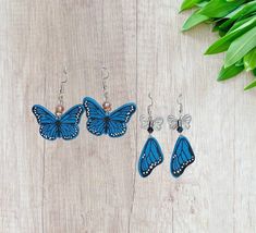 three pairs of earrings with blue butterflies on them sitting next to green leaves and flowers