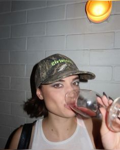 a woman wearing a camo cap drinking from a wine glass in front of a brick wall