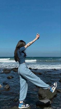 a woman standing on rocks near the ocean with her arms in the air and one leg up