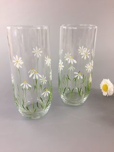 two glasses with daisies painted on them next to a white flower and green stems