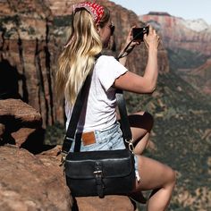 a woman sitting on top of a mountain taking a photo with her cell phone and camera