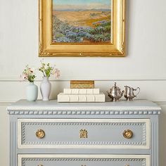 a painting hanging on the wall above a dresser with flowers and books in vases