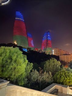 some very tall buildings lit up in the night sky with trees and bushes around them