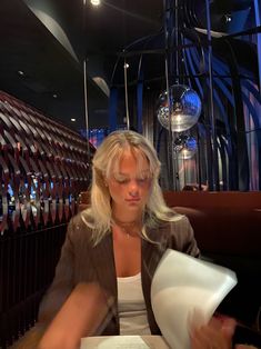 a woman sitting at a table reading a book and holding a piece of paper in her hand