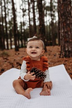 a baby is sitting on a blanket in the woods holding a pine cone and smiling