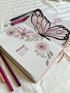 an open notebook with pink flowers and butterflies on it next to colored pencils, markers and a planner