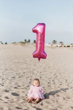 a baby sitting in the sand with a pink number one balloon