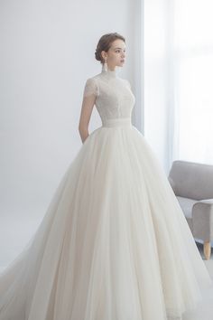 a woman in a white wedding dress is standing by a window and looking off to the side