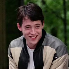 a smiling young man wearing a jacket and white t - shirt is sitting in front of trees