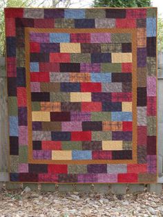 a multicolored quilt hanging on a wooden fence in front of some trees and bushes