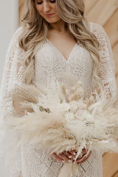 a woman wearing a white wedding dress holding a bouquet of dried flowers and feathers in her hands