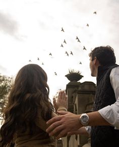 a man and woman standing next to each other with birds flying overhead in the sky