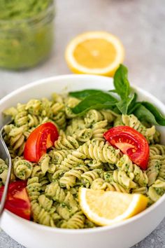 a white bowl filled with pesto pasta and topped with tomatoes, lemon wedges and basil