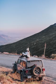 a man sitting on top of a black jeep in the middle of a mountain road