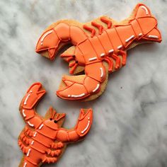 two cookies decorated to look like lobsters on a marble counter top, one is orange and the other has white icing