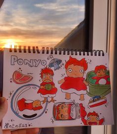 a person holding up a notebook with cartoon drawings on the front and back cover, in front of a window