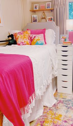 a white bed topped with pink and yellow pillows next to a dresser filled with books