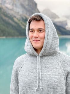 a man wearing a gray hoodie standing in front of a lake with mountains behind him