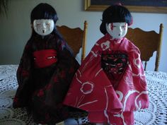 two dolls sitting on top of a table covered in red and black cloths next to each other