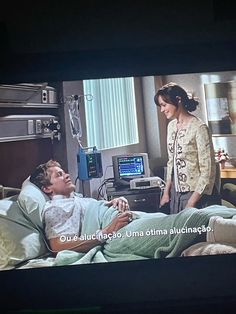 a woman standing next to a man laying in a hospital bed on a tv screen