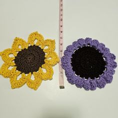 two crocheted flowers are next to a measuring tape on a table top, one is purple and the other is yellow