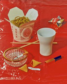 a red table topped with bowls and cups filled with noodles next to chopsticks