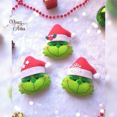 three green stuffed animals wearing santa hats on a white table with red beads and ornaments