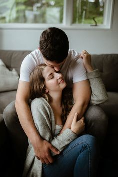 a man and woman cuddling on the couch in front of a window with their arms around each other