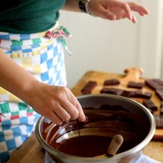 a woman mixing chocolate in a bowl on top of a wooden table with other food items