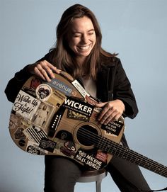 a woman sitting on a stool holding an acoustic guitar with stickers all over it