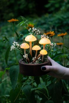 a hand holding a pot with mushrooms growing out of it in the middle of flowers