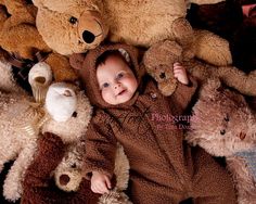 a baby in a bear suit surrounded by teddy bears and other stuffed animals, looking up at the camera