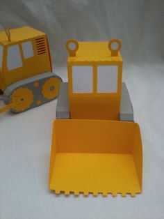 two construction vehicles made out of paper on a white tablecloth with a gray and yellow object in the background