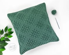 a green crocheted pillow next to a pair of knitting needles and a plant